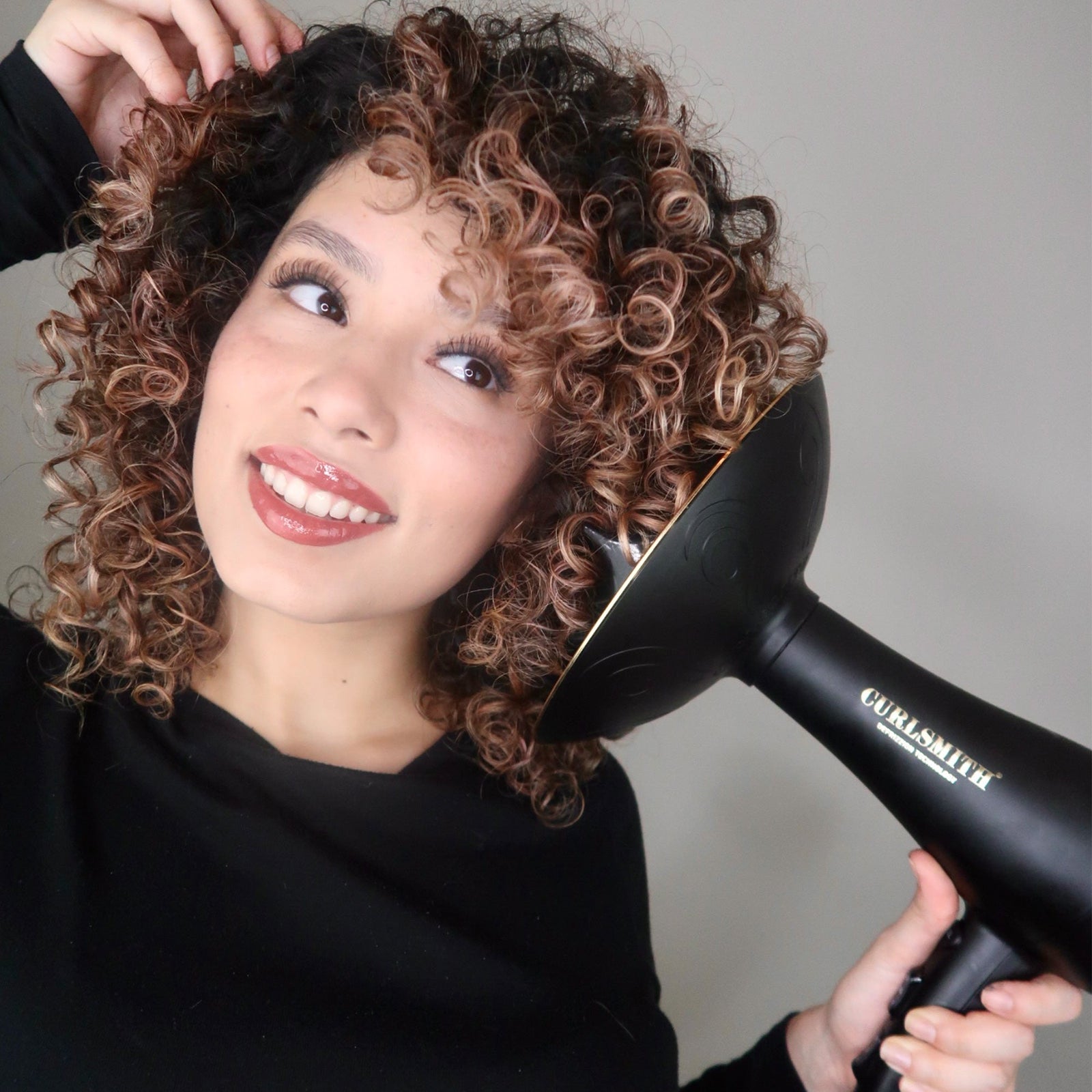 Woman using a hair dryer to style her curly hair against a neutral background