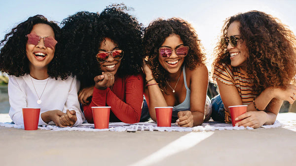 Four women with sunglasses and red cups sitting together outdoors.