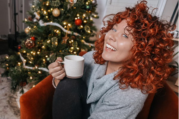 Woman with red curly hair holding a mug in front of a decorated Christmas tree.