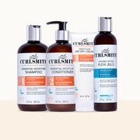Four Curlsmith hair care products arranged against a light cream background: two amber bottles with silver pumps (shampoo and conditioner), one white tube of air dry cream, and one dark amber bottle with a silver cap and blue label accent.