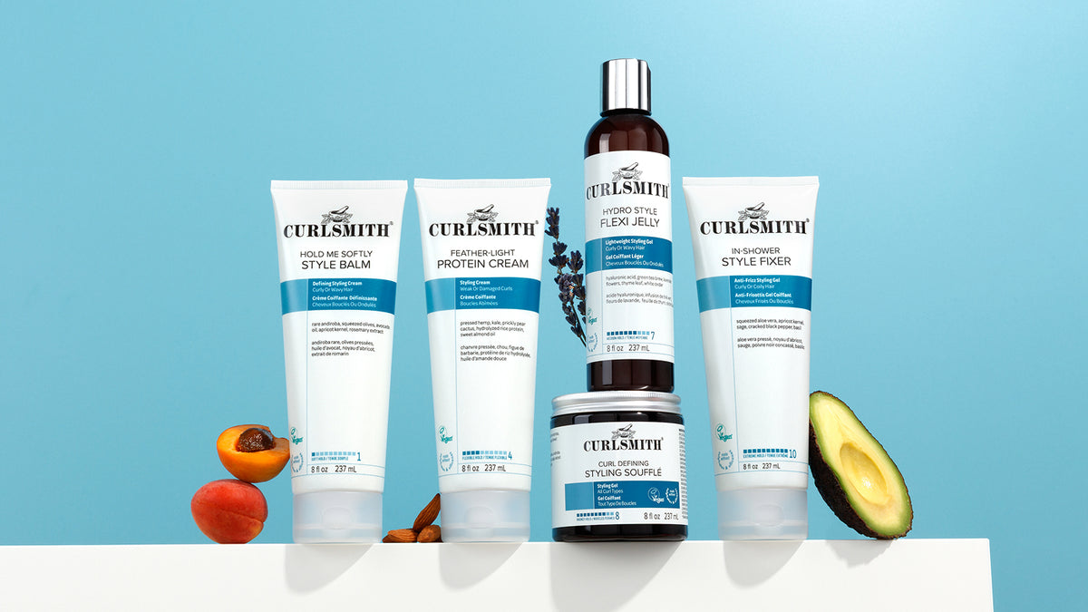 A set of Curlsmith hair care products arranged on a white surface against a light blue background, including Curlsmith's Hold Me Softly Style Balm, Feather Light Protein Cream, Hydro Style Flexi Jelly in a dark bottle, In-Shower Style Fixer, and Curl Defining Styling Souffle in a jar. Avocado and apricot slices are placed at the base beside the products.