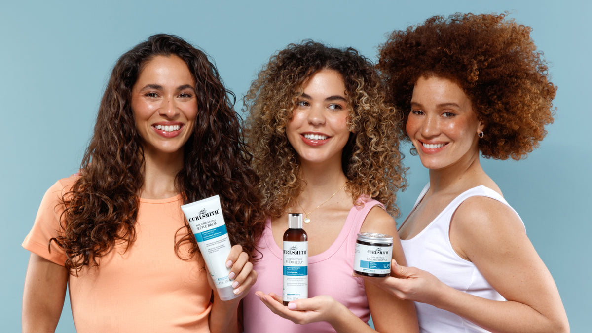 Three women with curly hair stand against a light blue background, each holding a skincare product from the brand Curlsmith. The products include Hold Me Softly Style Balm, a brown bottle of with white label called Hydro Style Flexi Jelly and a white jar with blue labels called Curl Defining Styling Souffle.