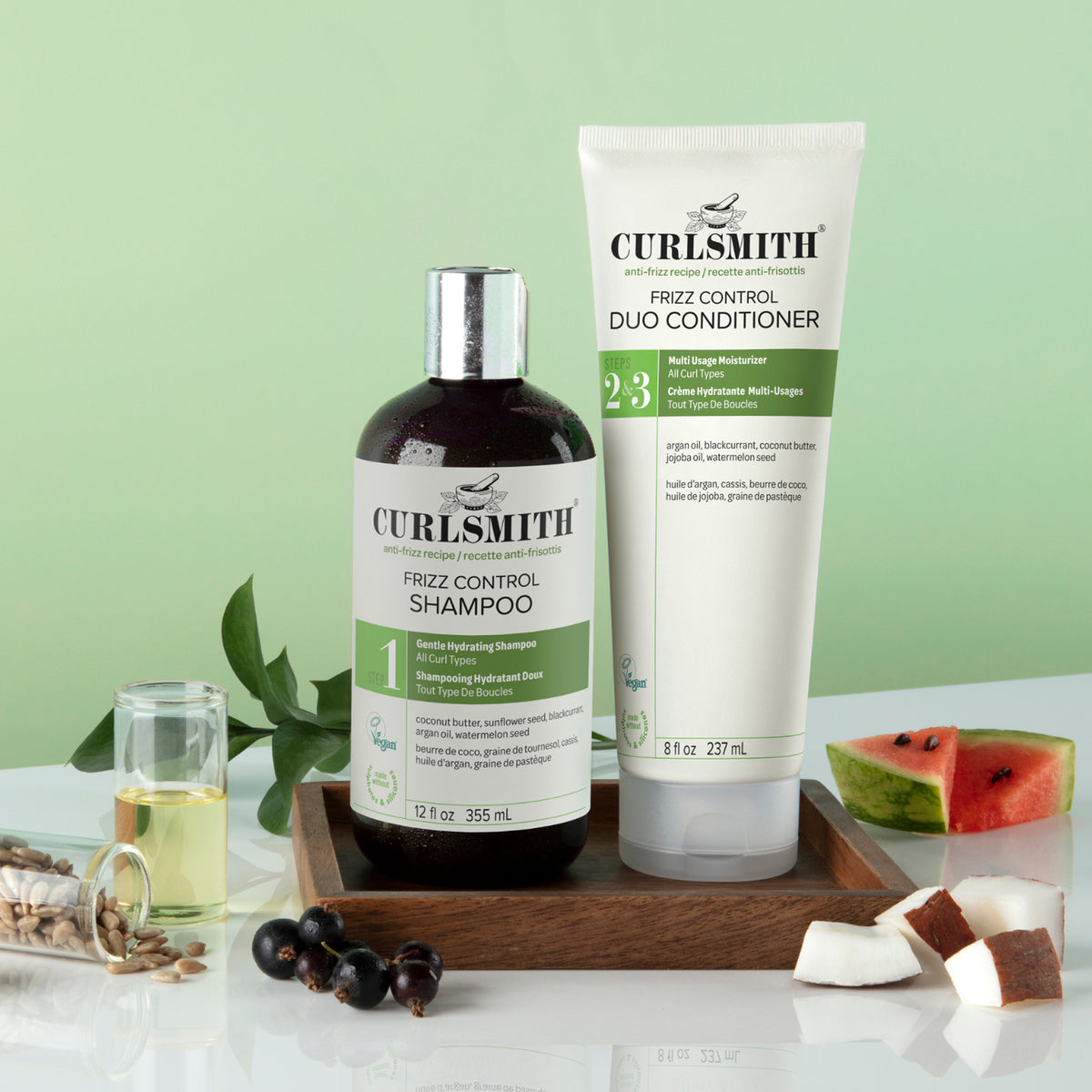 Curlsmith Frizz Control Duo set including a dark bottle of shampoo and a white conditioner tube, displayed on a wooden tray with coconut, watermelon, and botanical props against a light green background.
